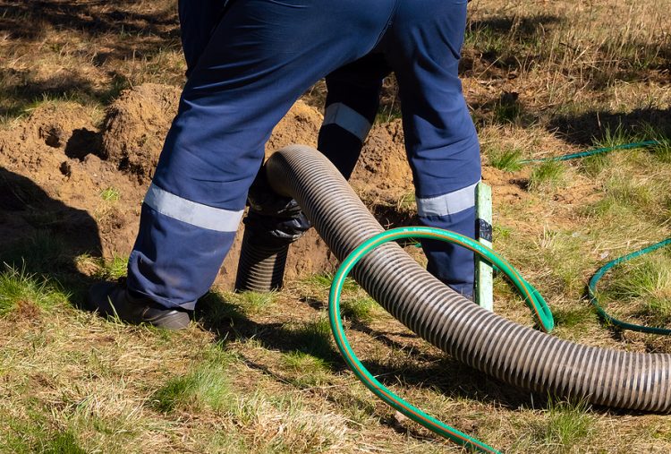 Man worker holding pipe, providing sewer cleaning service outdoor. Sewage pumping machine is unclogging blocked manhole